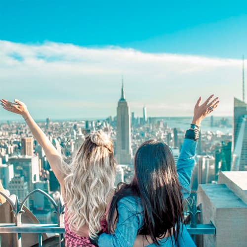 ~Two female friends on a New York highrise, arms in the air, looking out at the Empire State Building.